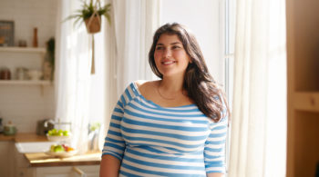 Beauty, bodypositivity, people and lifestyle concept. Indoor shot of gorgeous brunette Latin girl posing by window in kitchen dressed in xxl blue and white striped shirt, having joyful beaming smile