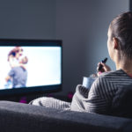 Woman watching romantic movie and eating ice cream. Sad lonely single girl streaming series or film on tv home at night. Person with stream on television.